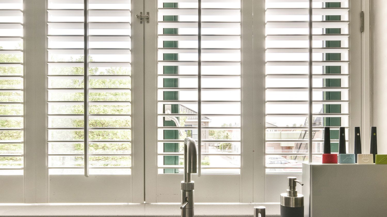 Crisp white plantation shutters above a kitchen sink, daylight and trees visible through the open louvres