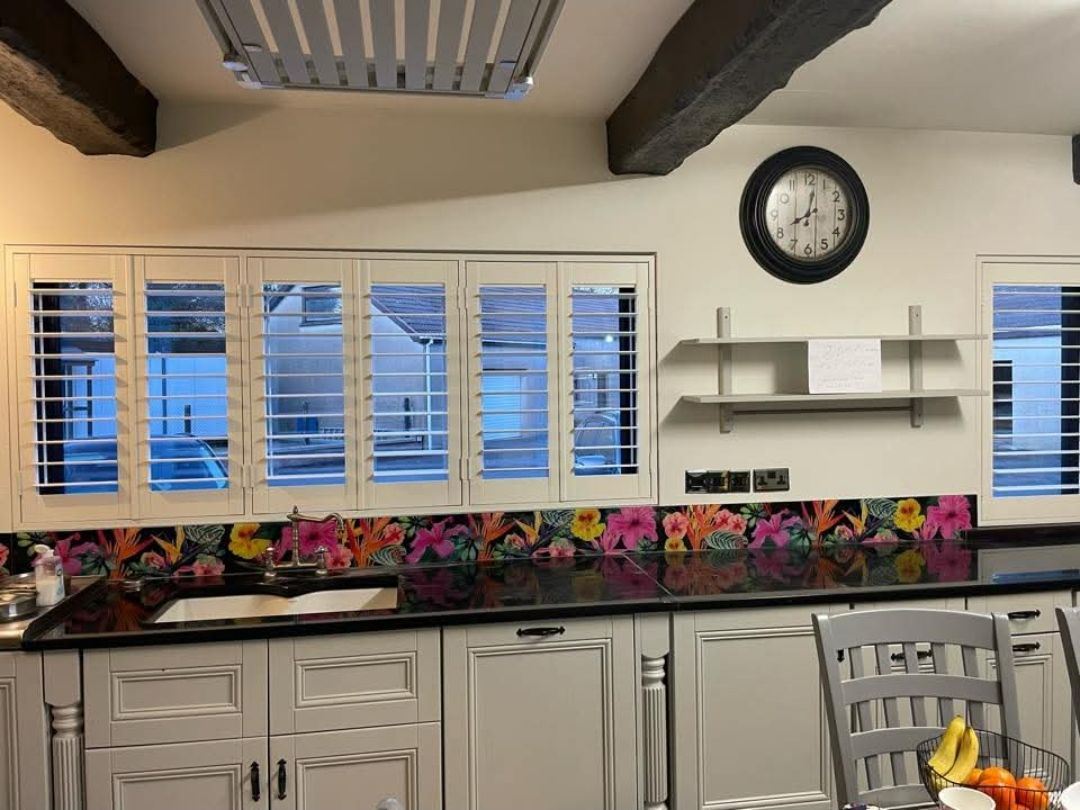 Cream full-height plantation shutters across five kitchen windows above a black granite worktop and floral splashback